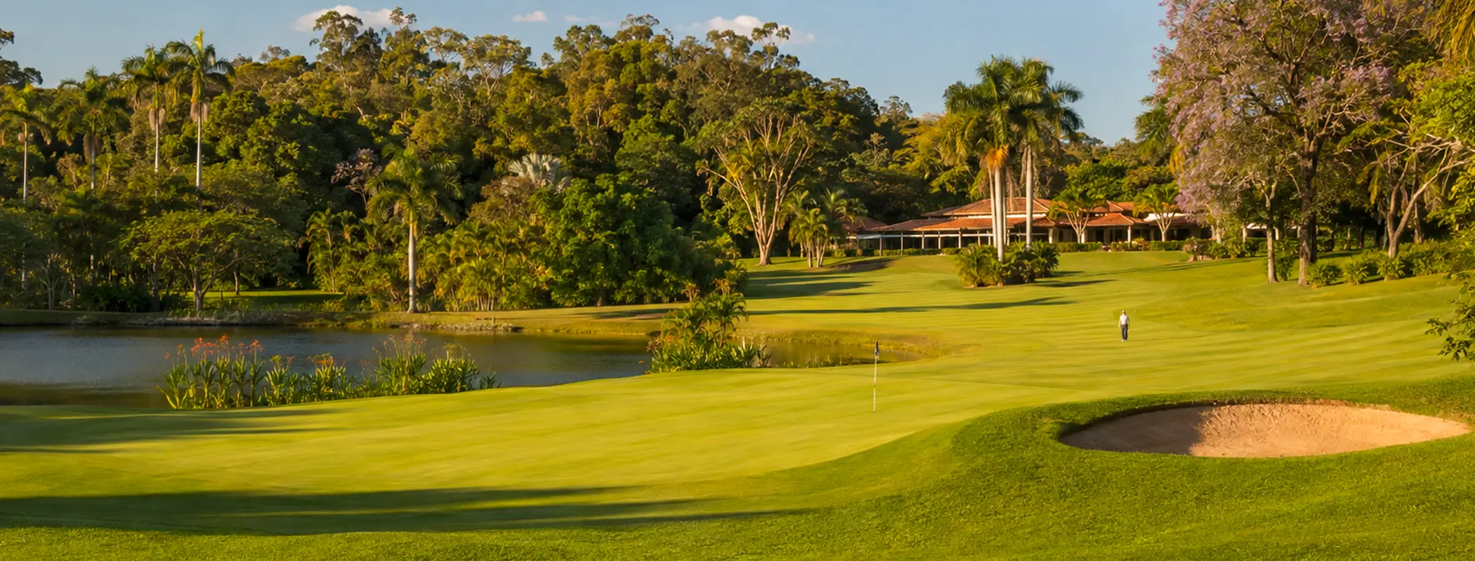 Vista panorâmica de um campo de golfe, com green, bunker de areia, lago e a sede do clube ao fundo