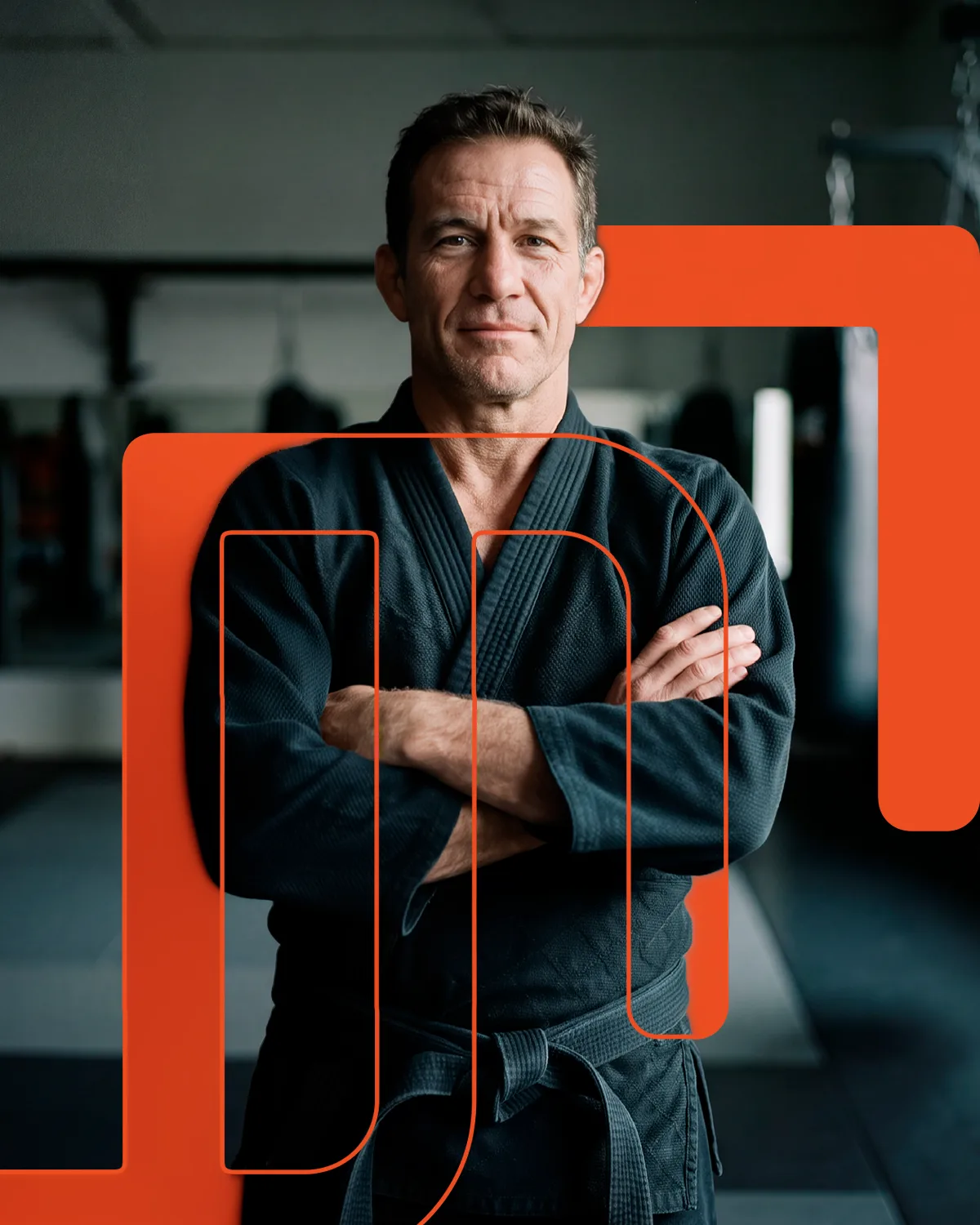 Professor de artes marciais de quimono escuro e braços cruzados, com os sacos de pancada da academia ao fundo